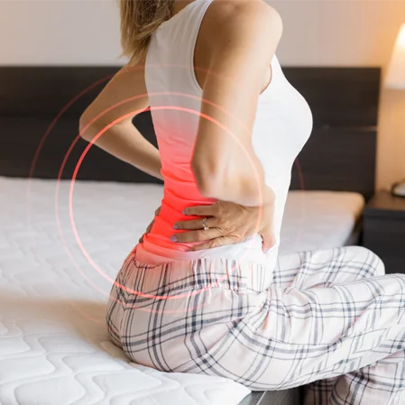 A woman sitting on the edge of the bed holds her back in pain. Red highlighting indicated the pain radiating from her back