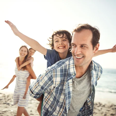 A little boy spreads his arms in joy from where he rides in a piggyback on his father's back. His family walks on the beach