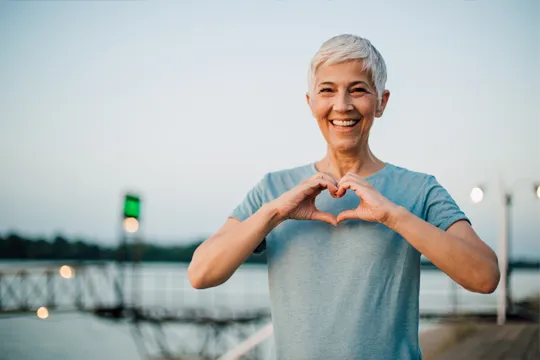 A woman standing on a pier on a cloudy day. She smiles and holds her hands up to create a heart shape.